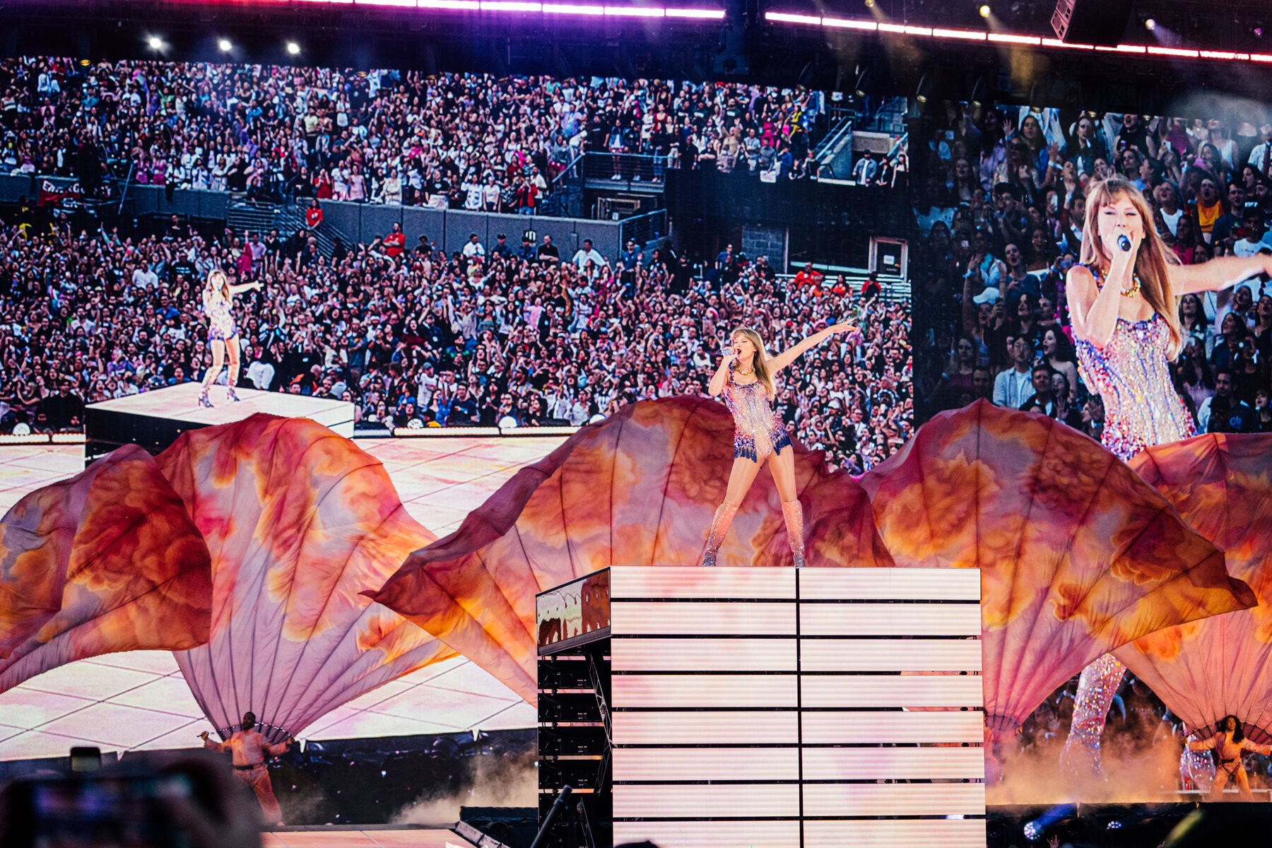 Taylor Swift, in a sparkly leotard, stands onstage on a riser while singing into a microphone with one arm extended in the air. On video screens behind her, the large crowd is visible.