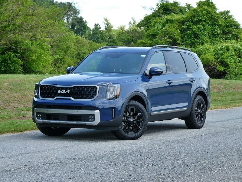 Front ¾ view of a 2023 Kia Telluride in dark green parked on a paved road in a suburban setting with houses and trees in the background