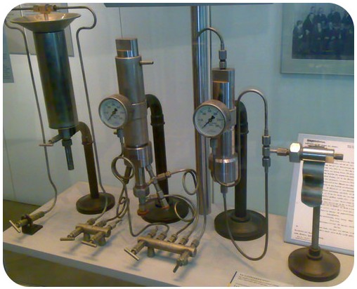 A display of vintage scientific instruments with pressure gauges and metal tubes on a tabletop. The setup includes various cylindrical components and dials, situated in a museum-like setting.