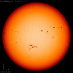 Large orange-red sphere which is the sun, against the black backdrop of space. Dark black patches are visible speckled across the surface of the sun, these are sunspots