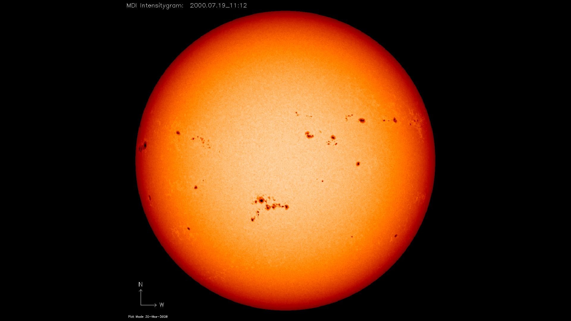 Large orange-red sphere which is the sun, against the black backdrop of space. Dark black patches are visible speckled across the surface of the sun, these are sunspots