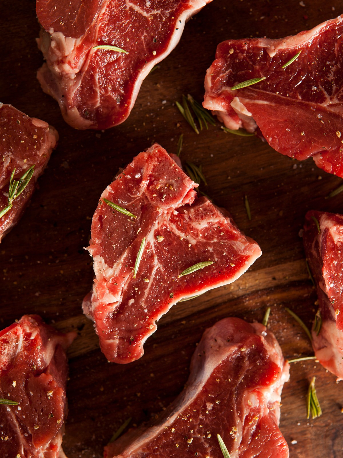 A close-up shot of seasoned, uncooked lamb loin chops displayed on a cutting board, looking fresh and flavorful with salt, pepper, and bits of rosemary on top of each.