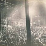 A large crowd gathers in the street at night under a lit up archway.