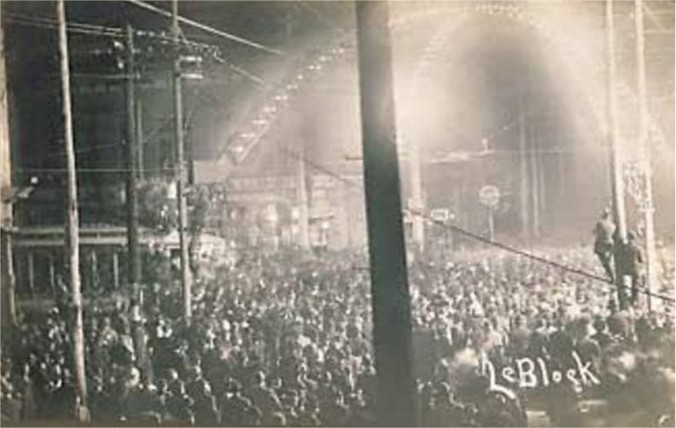 A large crowd gathers in the street at night under a lit up archway.