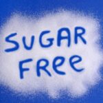 A blue background sprinkled with white sugar substitute crystals with the words sugar free written; concept is sugar alcohols and artficial sweeteners