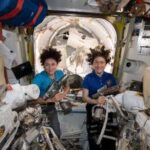 Two female astronauts in the international space station behind spacesuits
