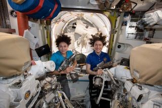 Two female astronauts in the international space station behind spacesuits