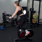 Woman standing up riding a Peloton alternative, the echelon ex5 bike with other gym equipment in the background