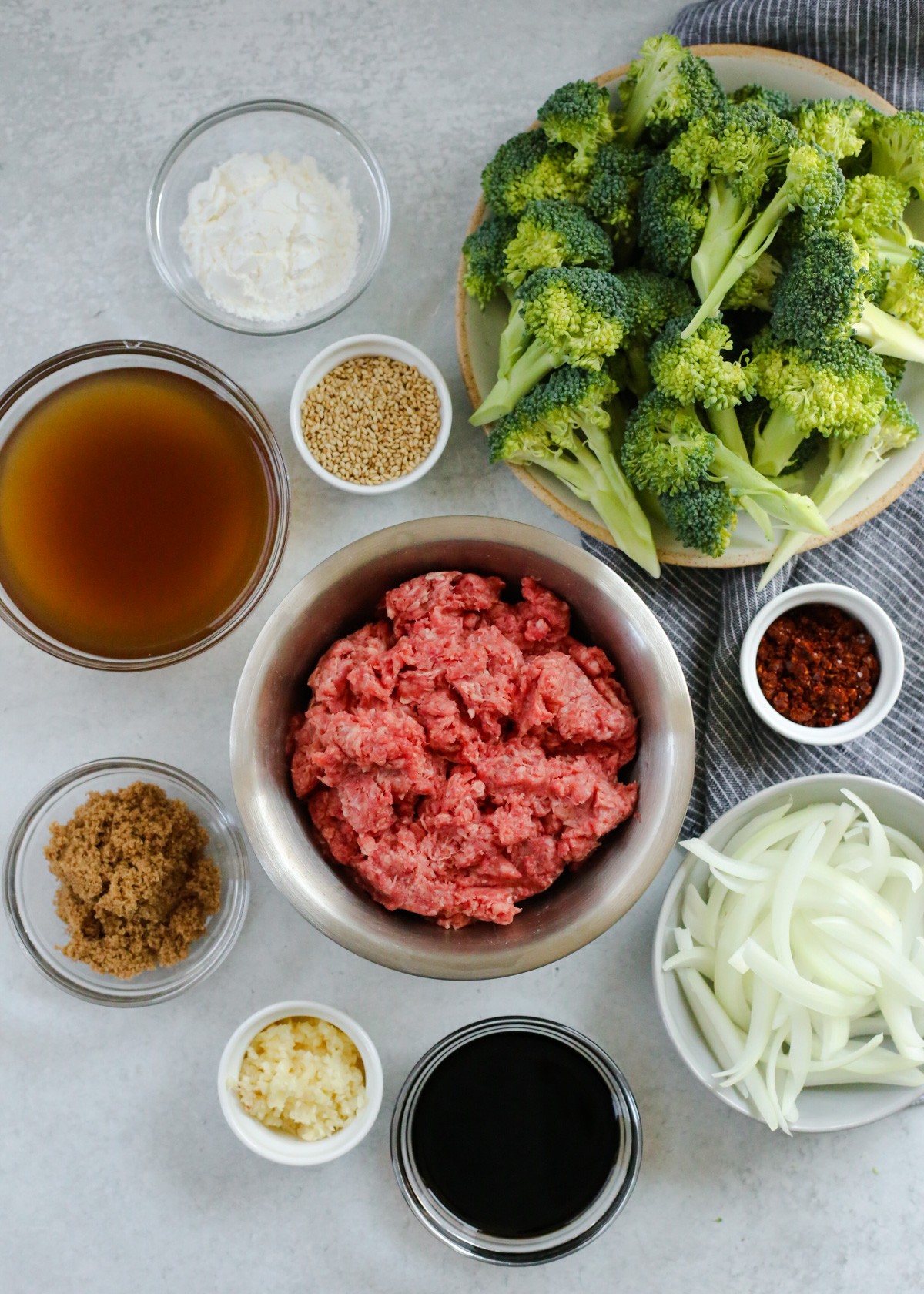 A small silver prep bowl filled with uncooked ground beef is shown next to the other ingredients needed for a Beef and Broccoli recipe.